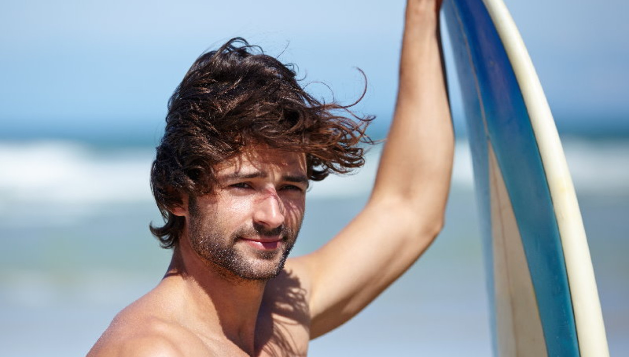 attraktiver Mann am Strand, der ein Surfbrett hält