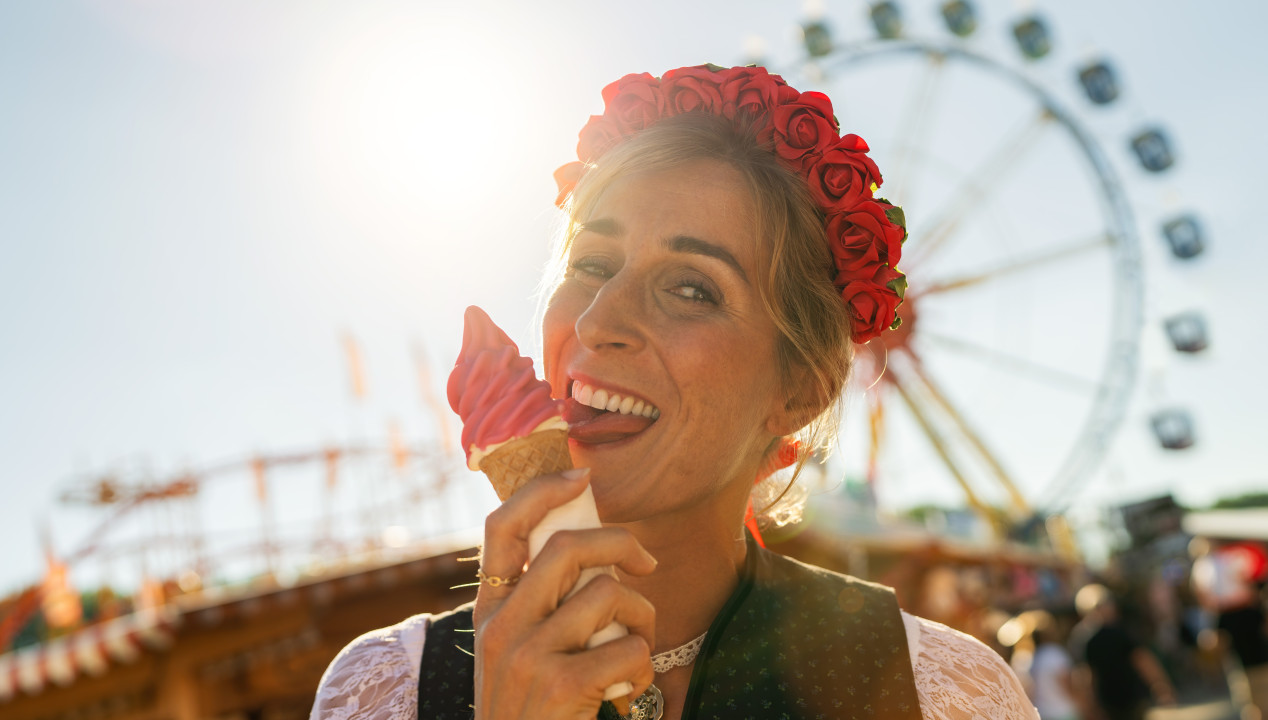 Attraktive Frau steht im Dirndl auf dem Oktoberfest vor dem Riesenrad und lutscht ein Eis